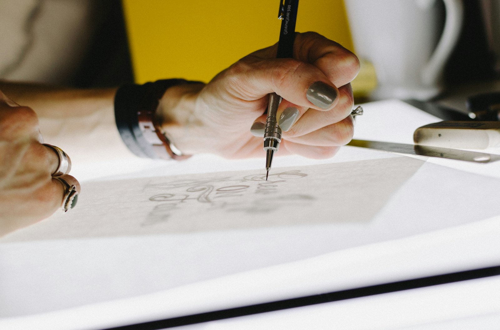 A hand practicing deliberate lettering with a mechanical pencil on tracing paper, using a ruler for precision